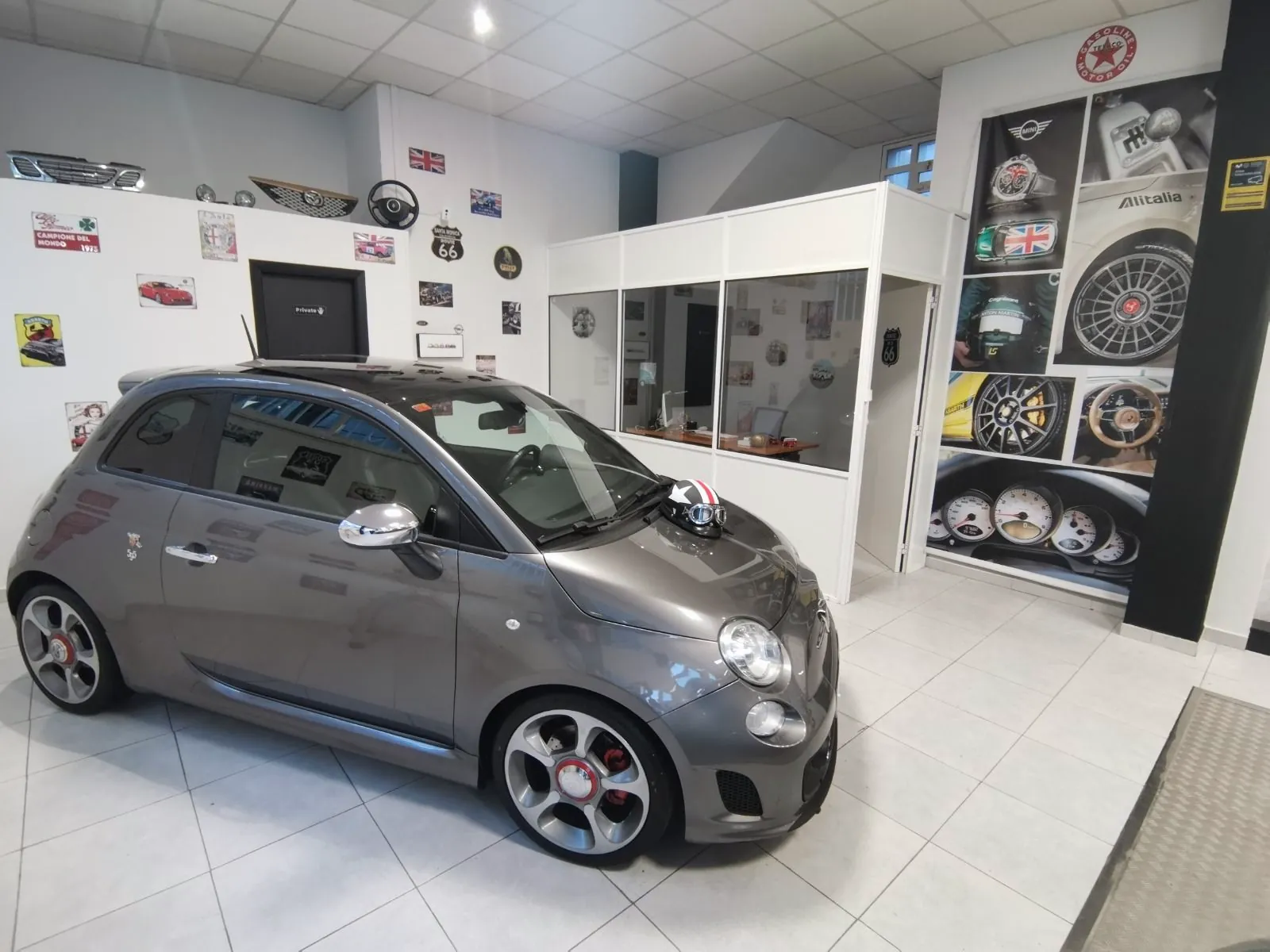 Fiat 500 Abarth gris en el local de Brigam Cars, entre paredes decoradas con placas y carteles de motor.