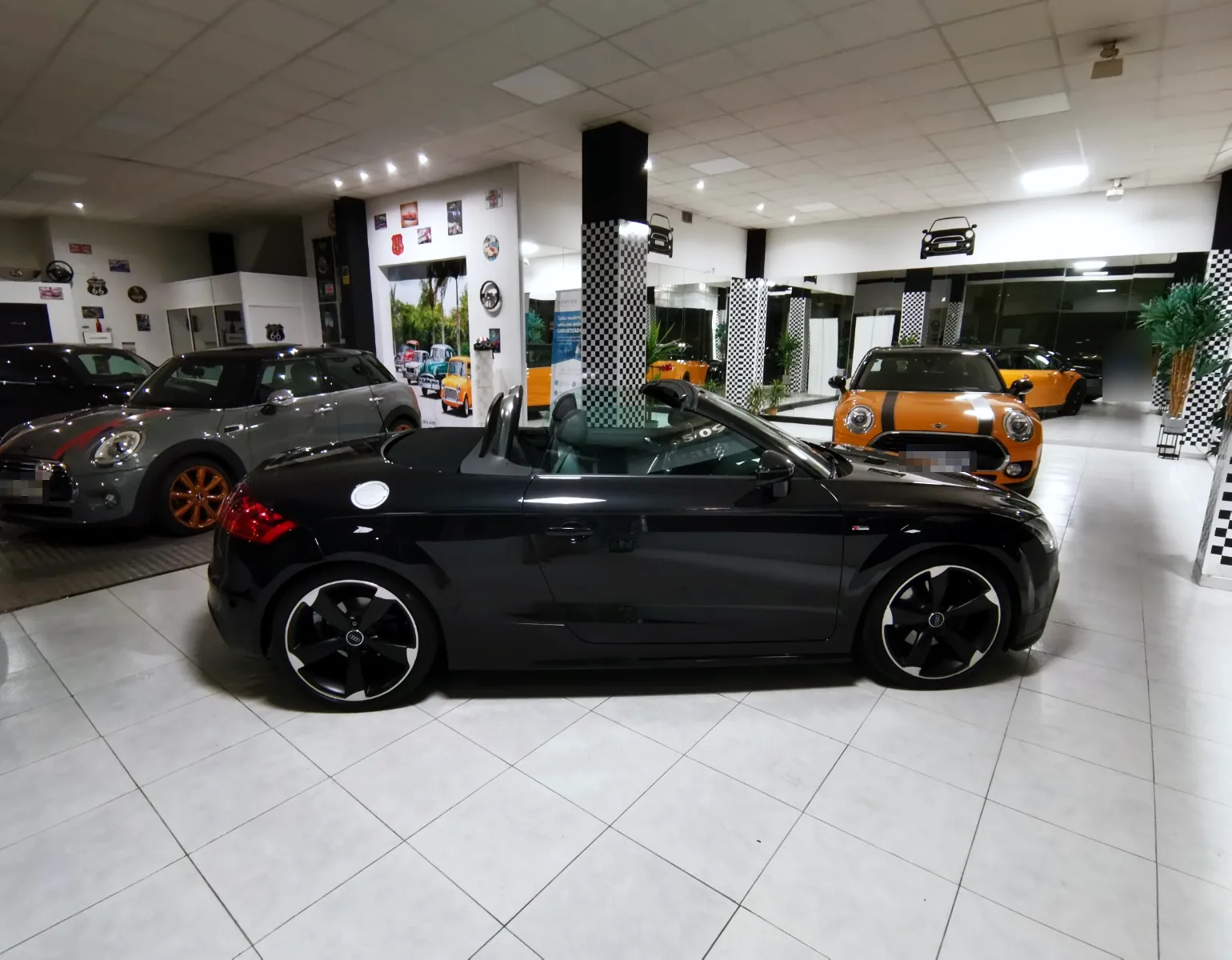 Vista general del salón de Brigam Cars: un Audi TT descapotable negro en el centro, dos Mini al fondo, columnas de cuadros blancos y negros y una pared de espejos.
