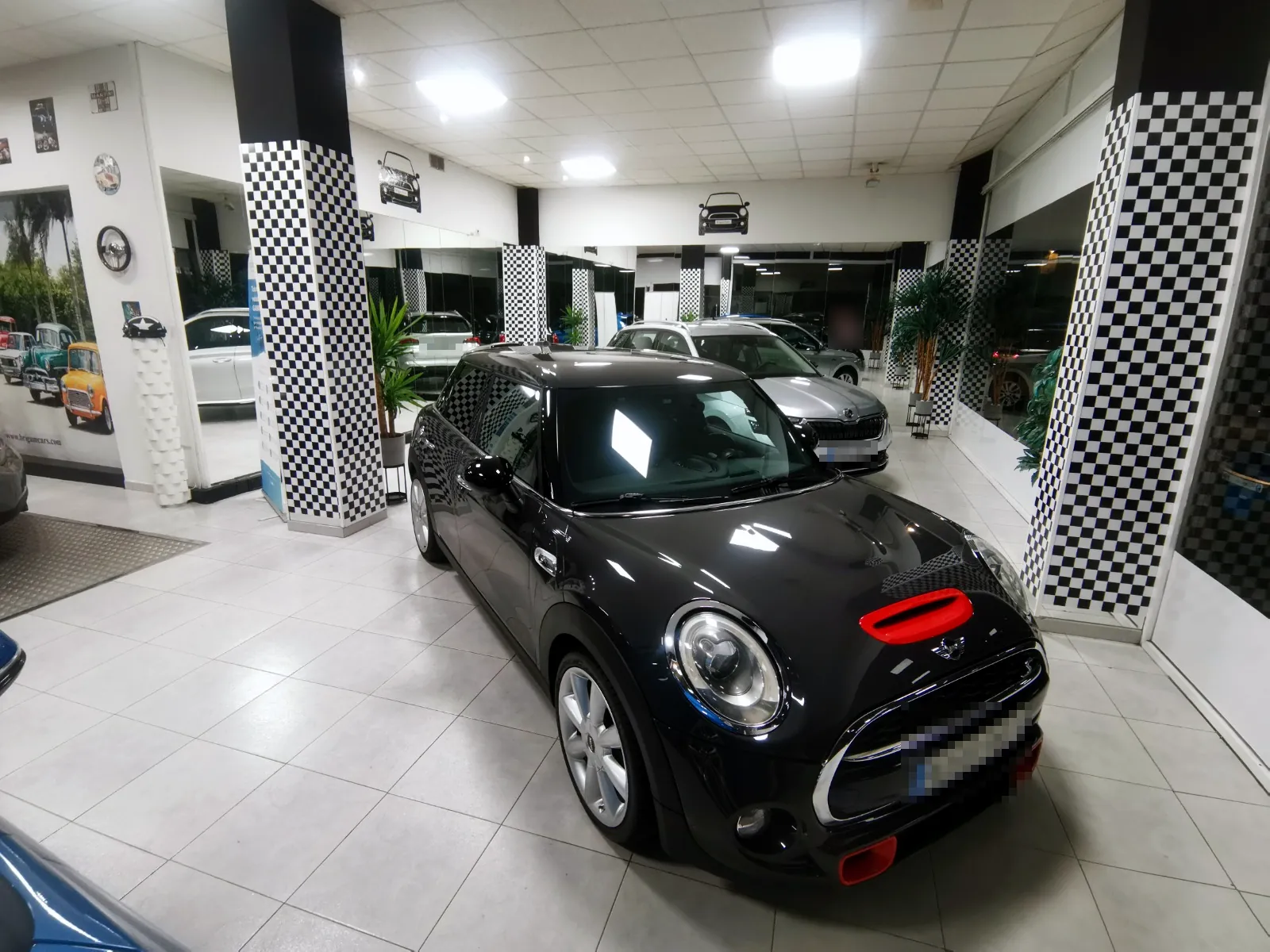 Mini negro en primer plano en el salón de Brigam Cars, con un Skoda blanco detrás, entre columnas de cuadros blancos y negros.