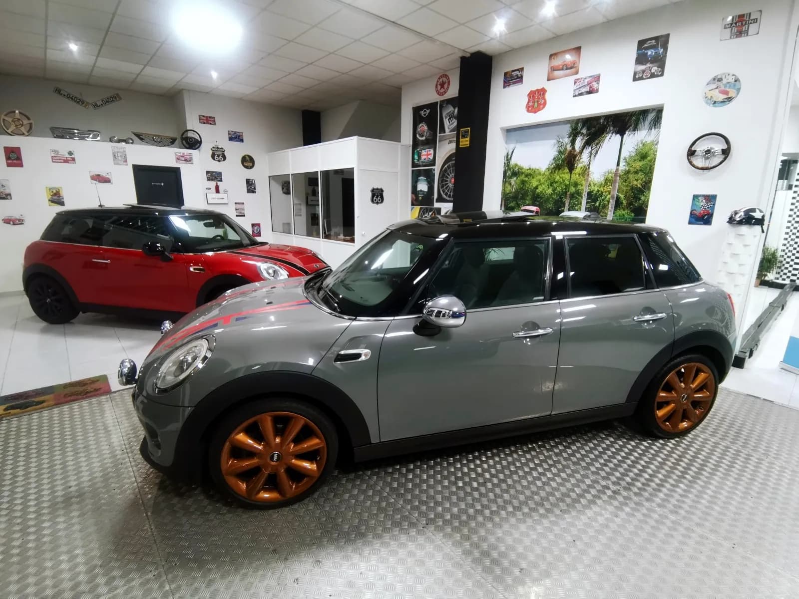Mini gris de cinco puertas con llantas naranjas sobre la plataforma del salón de Brigam Cars; detrás, un Mini rojo y la pared decorada con placas de motor.
