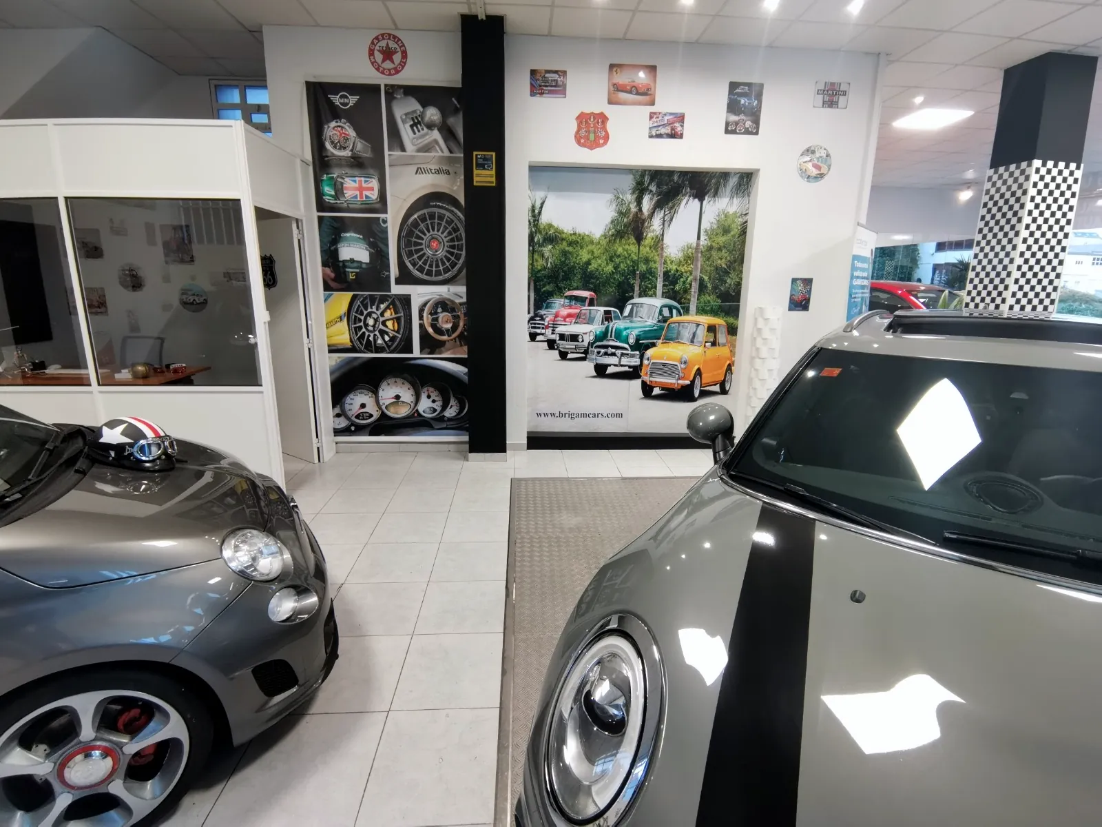Rincón del salón de Brigam Cars con un fotomural de coches clásicos y placas de motor en la pared, entre un Fiat 500 Abarth y un Mini grises.
